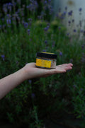 A hand holding a small jar of honey outdoors in front of a blurred background of purple lavender flowers. The jar has a black lid and a yellow label with a honeycomb design.
