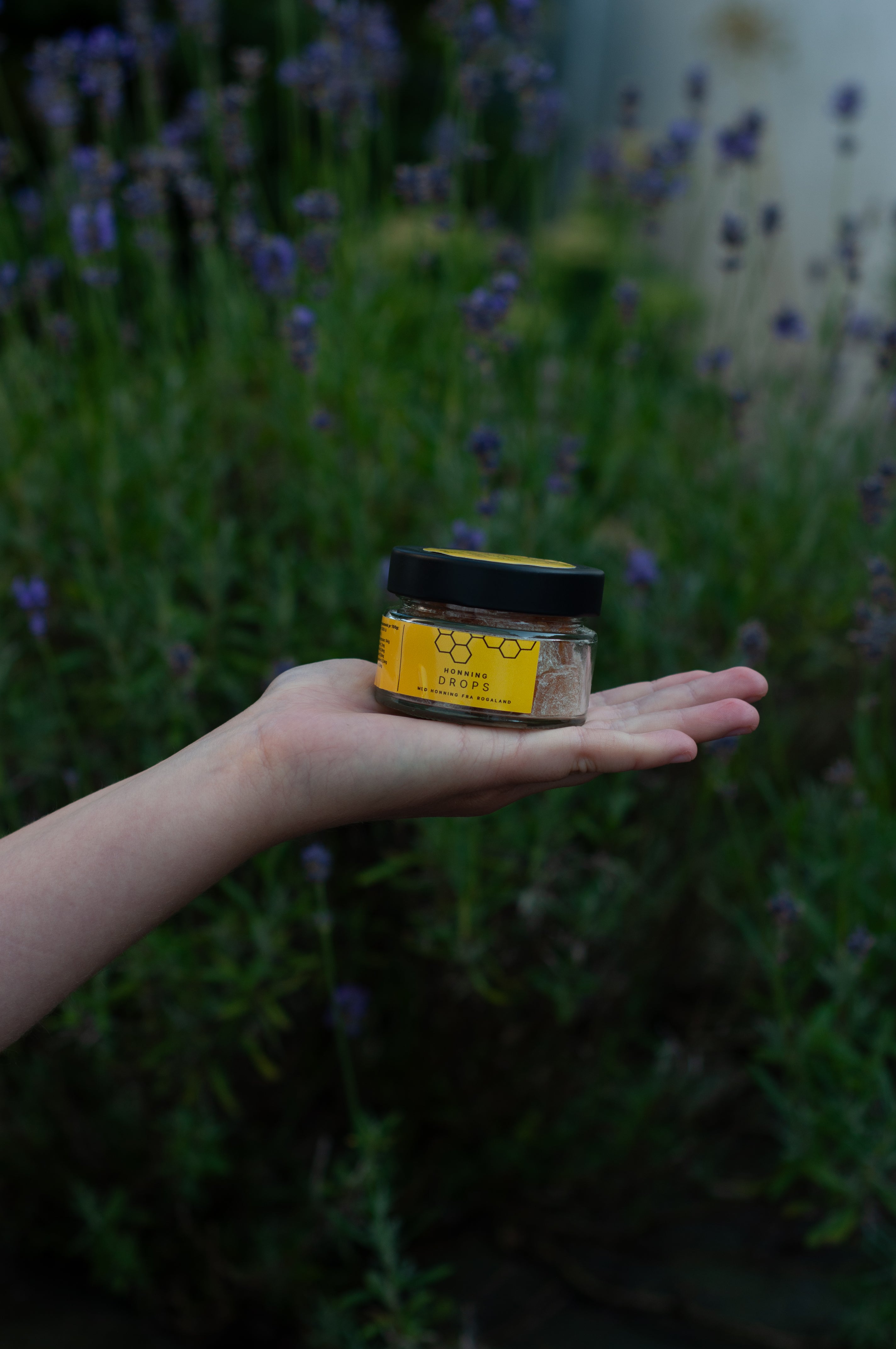 A hand holding a small jar of honey outdoors in front of a blurred background of purple lavender flowers. The jar has a black lid and a yellow label with a honeycomb design.